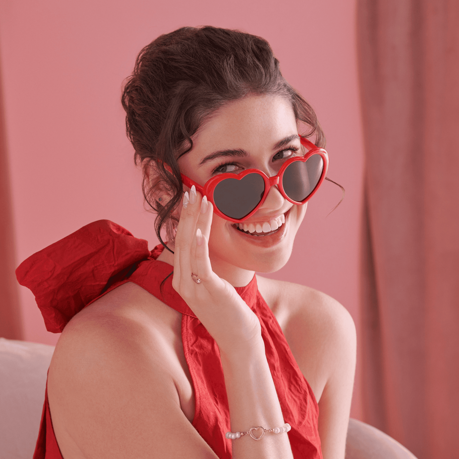 Smiling woman in a red halter dress and red heart-shaped sunglasses shows off white press-on nails with heart designs on a pink background.