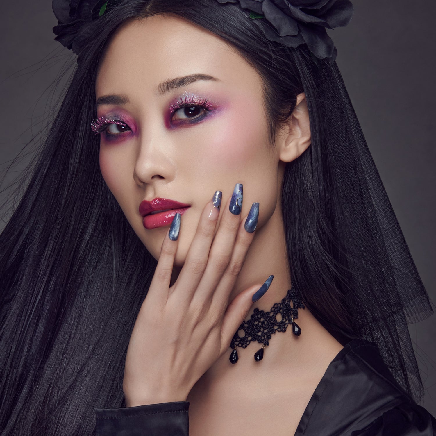 Close-up of a woman with long black hair wearing sparkly pink false eyelashes decorated for Halloween, vibrant purple eyeshadow, glossy dark pink lips, marbled blue nails with moon and star details, a black lace choker, and a black floral headpiece with a veil.