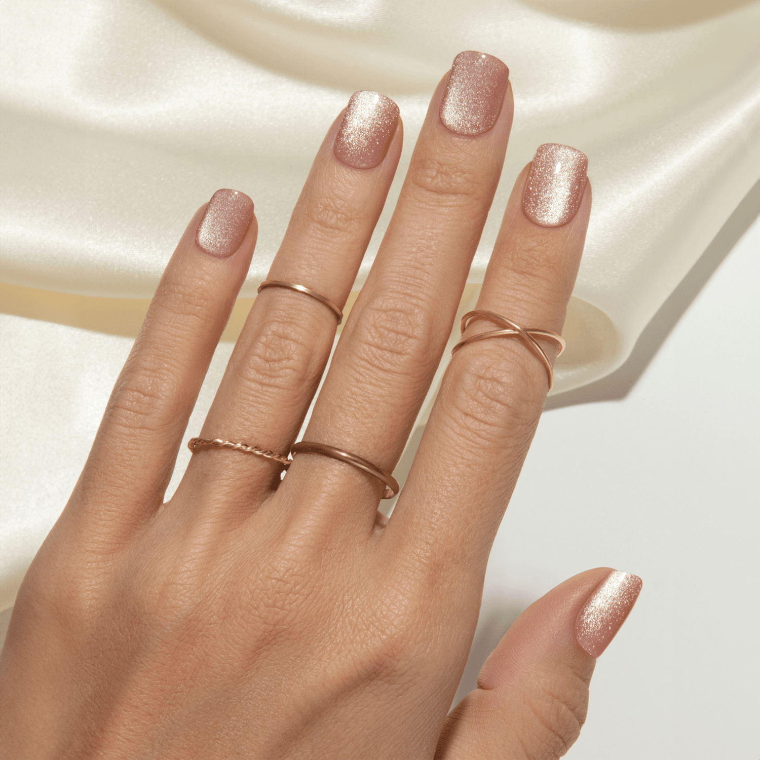 Hand with light to medium skin tone wearing glittery rose gold press-on nails in a square shape, styled with thin gold rings on a silky cream background.
