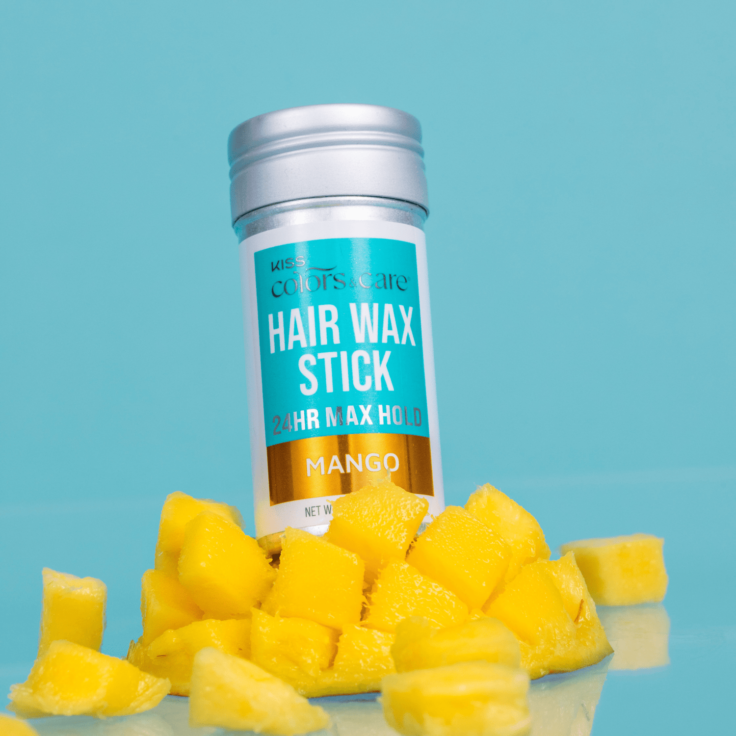 Silver-capped KISS Colors &amp; Care Hair Wax Stick with blue, white, and gold label, surrounded by fresh yellow mango chunks on blue background.