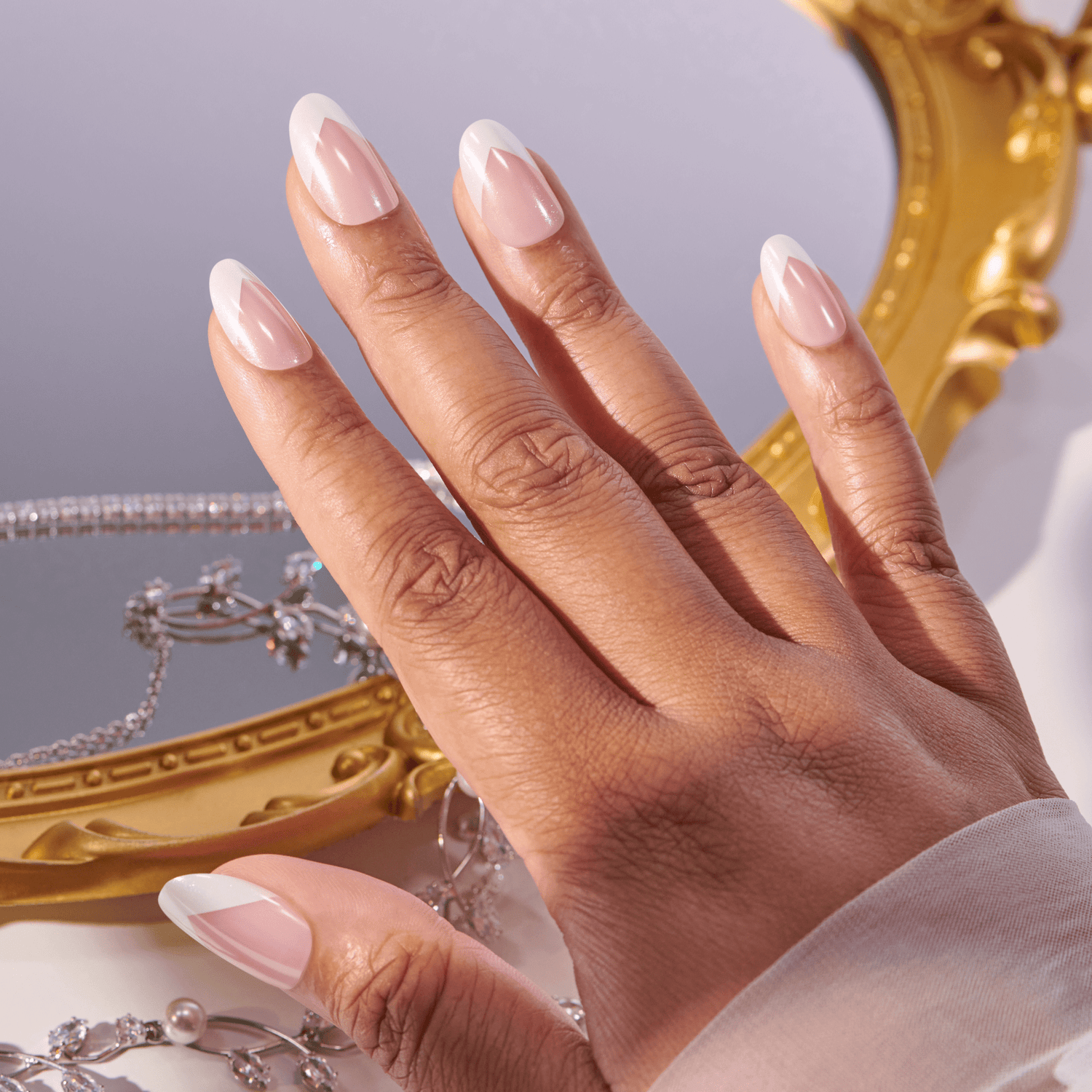 Hand with medium brown skin showing almond-shaped nails painted pink with white geometric French tips, posed over a reflective surface with jewelry and a gold-framed mirror.