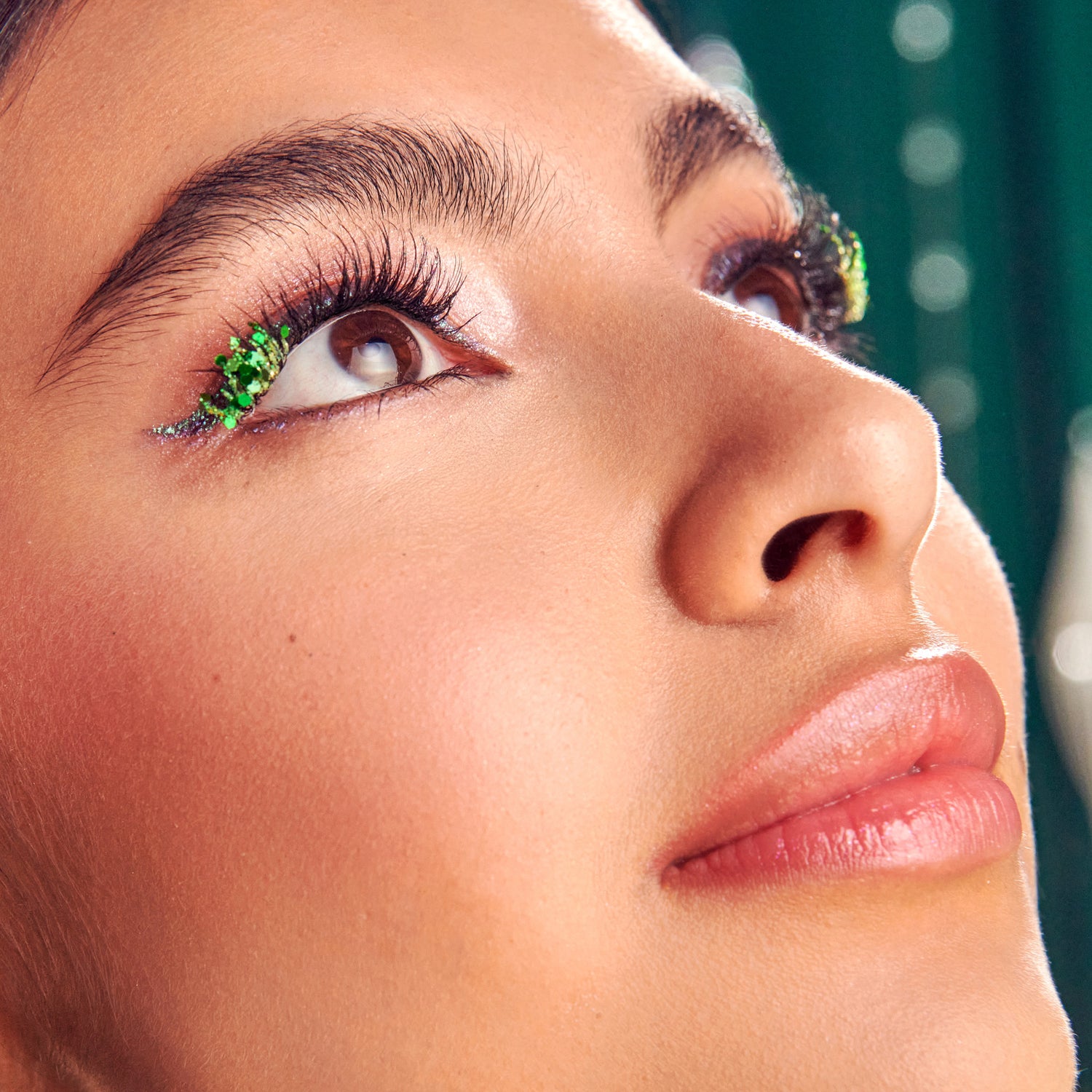 Close-up of eye makeup with bright green glitter and long black lashes against teal background.