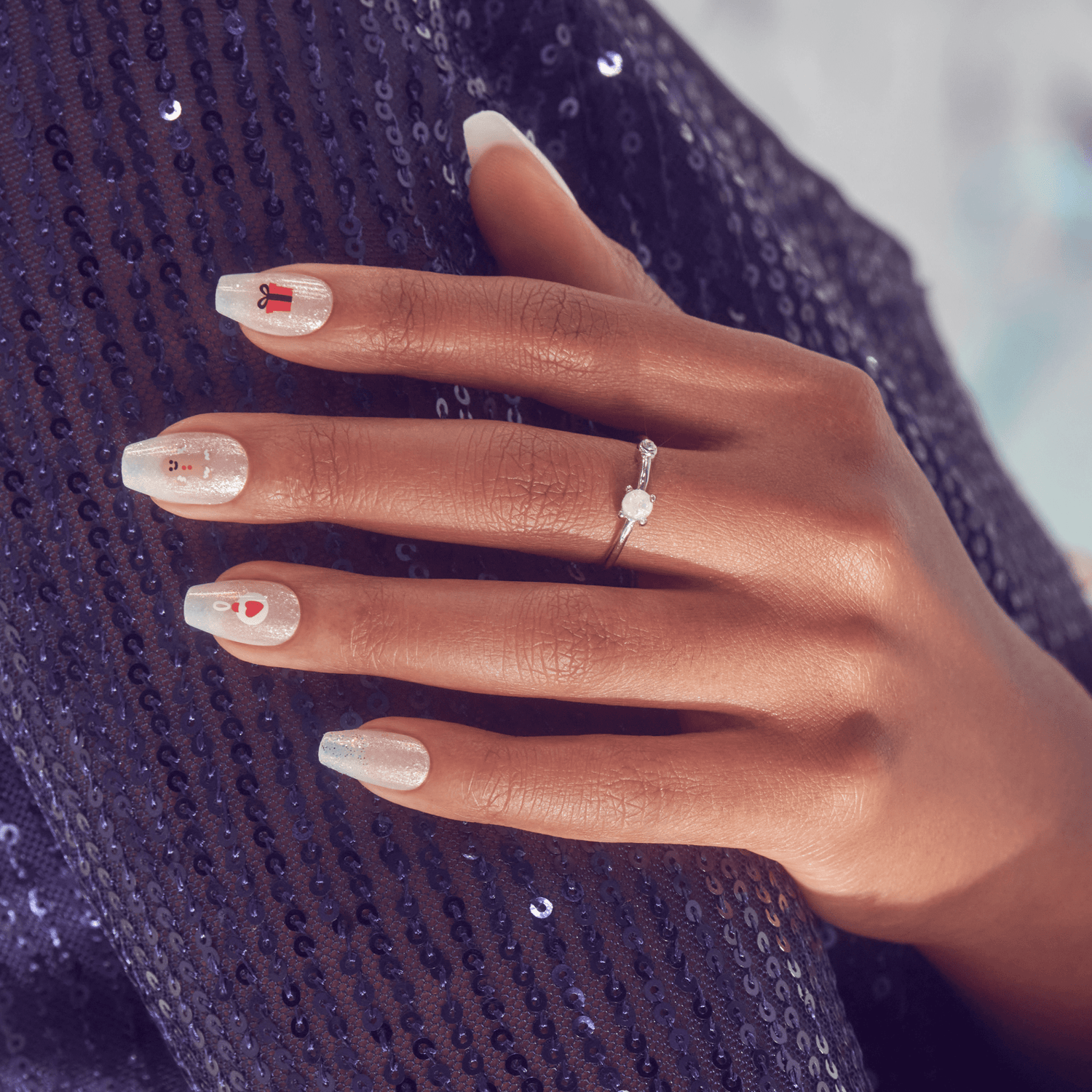 Hand with medium brown skin shows white, shimmery acrylic nails decorated with small holiday designs including a red gift box, snowman, and candy cane, resting on dark blue sequined fabric.