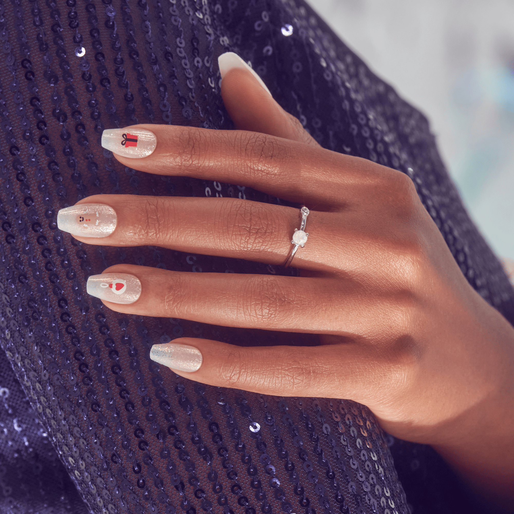 Hand with medium brown skin shows white, shimmery acrylic nails decorated with small holiday designs including a red gift box, snowman, and candy cane, resting on dark blue sequined fabric.