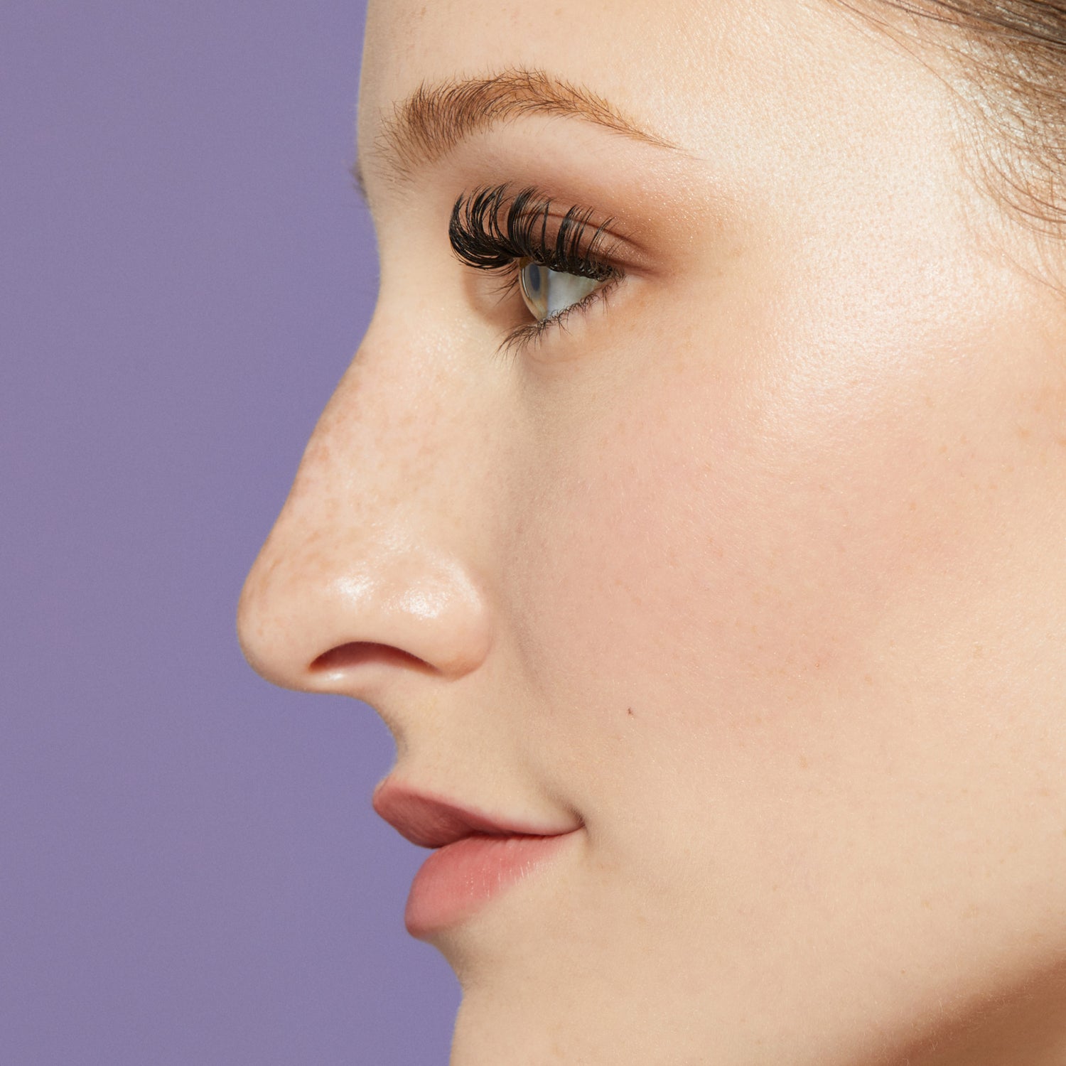 Close-up side profile of a person with natural skin and freckles wearing long, thick L curl false eyelashes against a purple background.
