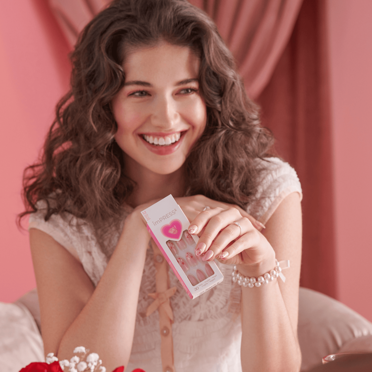 Smiling woman with curly brown hair wears pink-themed press-on nails with heart designs, holding imPRESS packaging showing the same nail style.