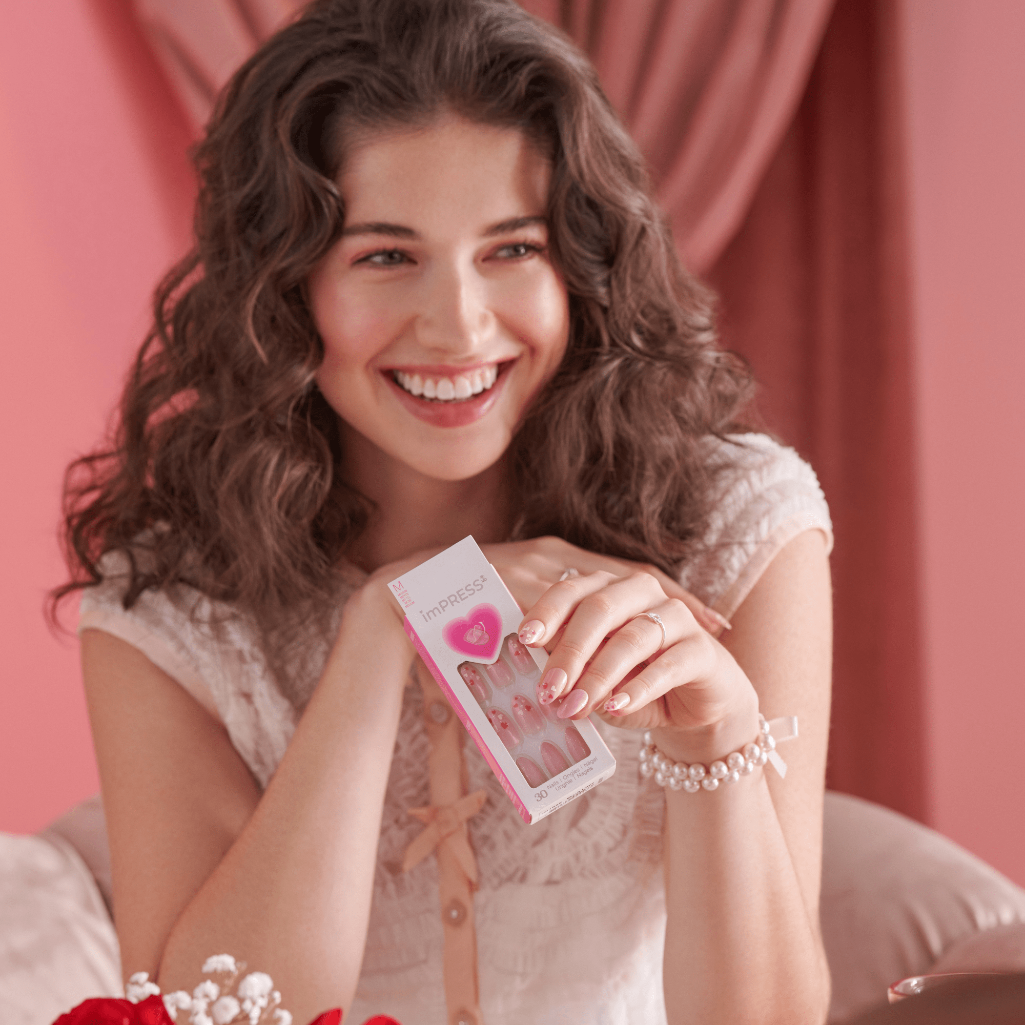Smiling woman with curly brown hair wears pink-themed press-on nails with heart designs, holding imPRESS packaging showing the same nail style.