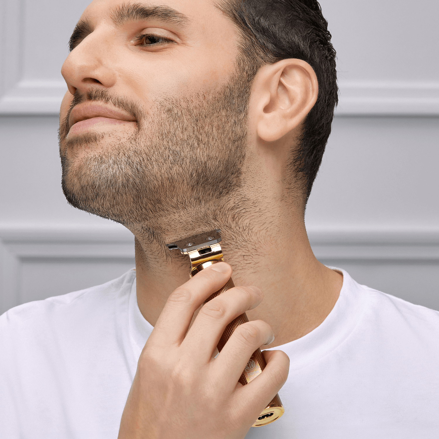 Man with short dark hair and beard wearing a white shirt trims his neck hair using a gold cordless precision blade trimmer against a gray paneled background.