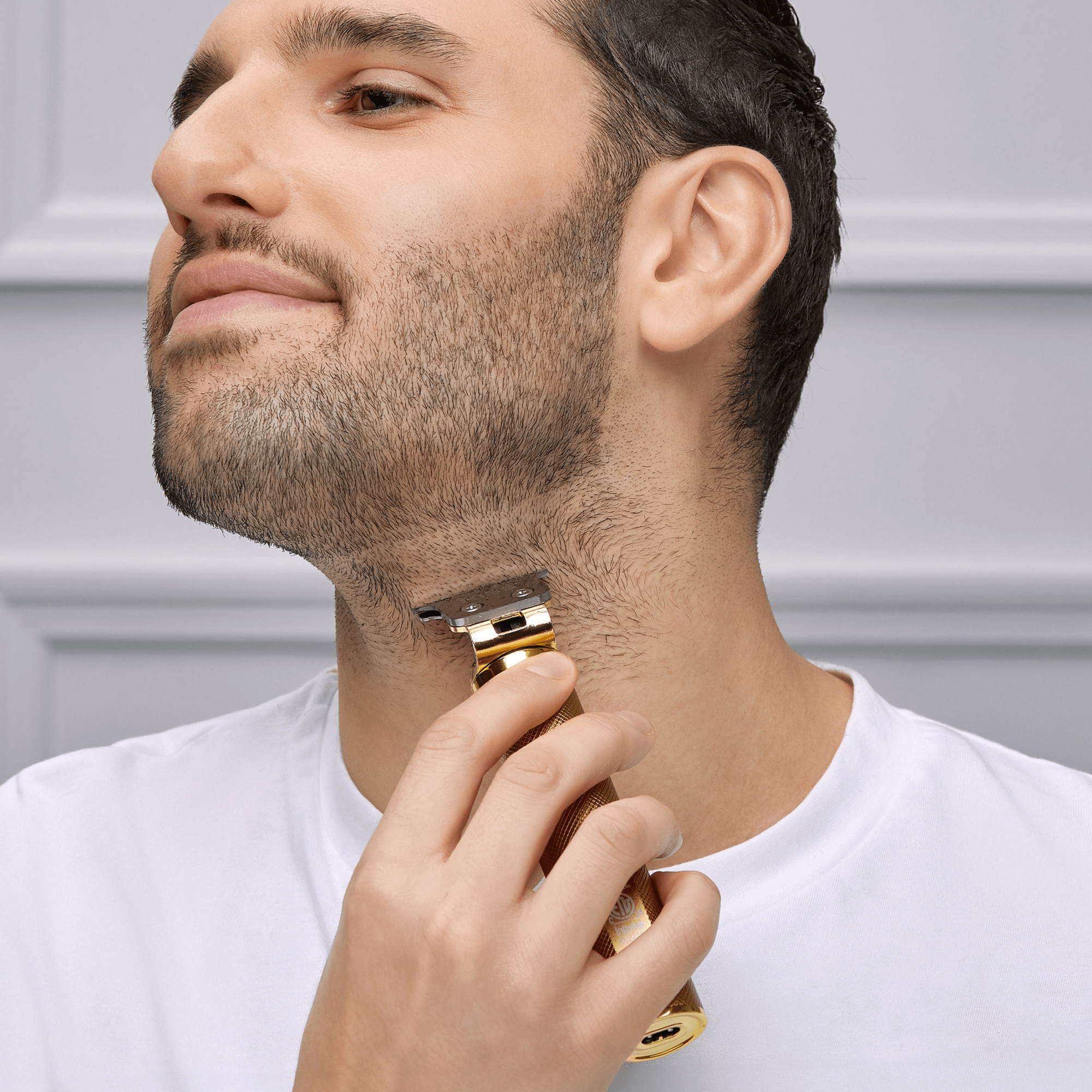 Man with short dark hair and beard wearing a white shirt trims his neck hair using a gold cordless precision blade trimmer against a gray paneled background.