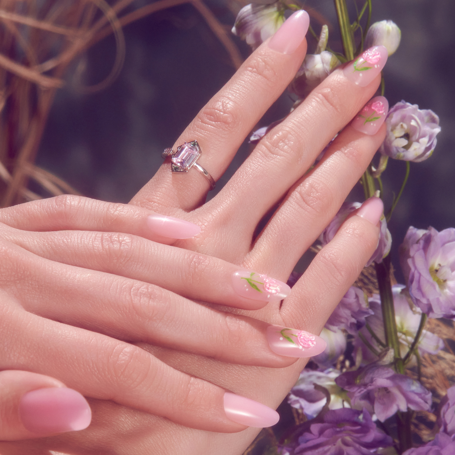 Hands with soft pink almond-shaped nails featuring delicate small pink rose designs on some nails, posed against light purple flowers.