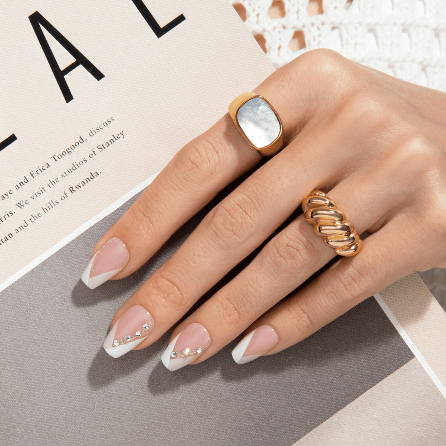 Hand with French press on nails in a soft pink shade with white angled tips and small rhinestone accents, resting on a beige and gray textured surface.