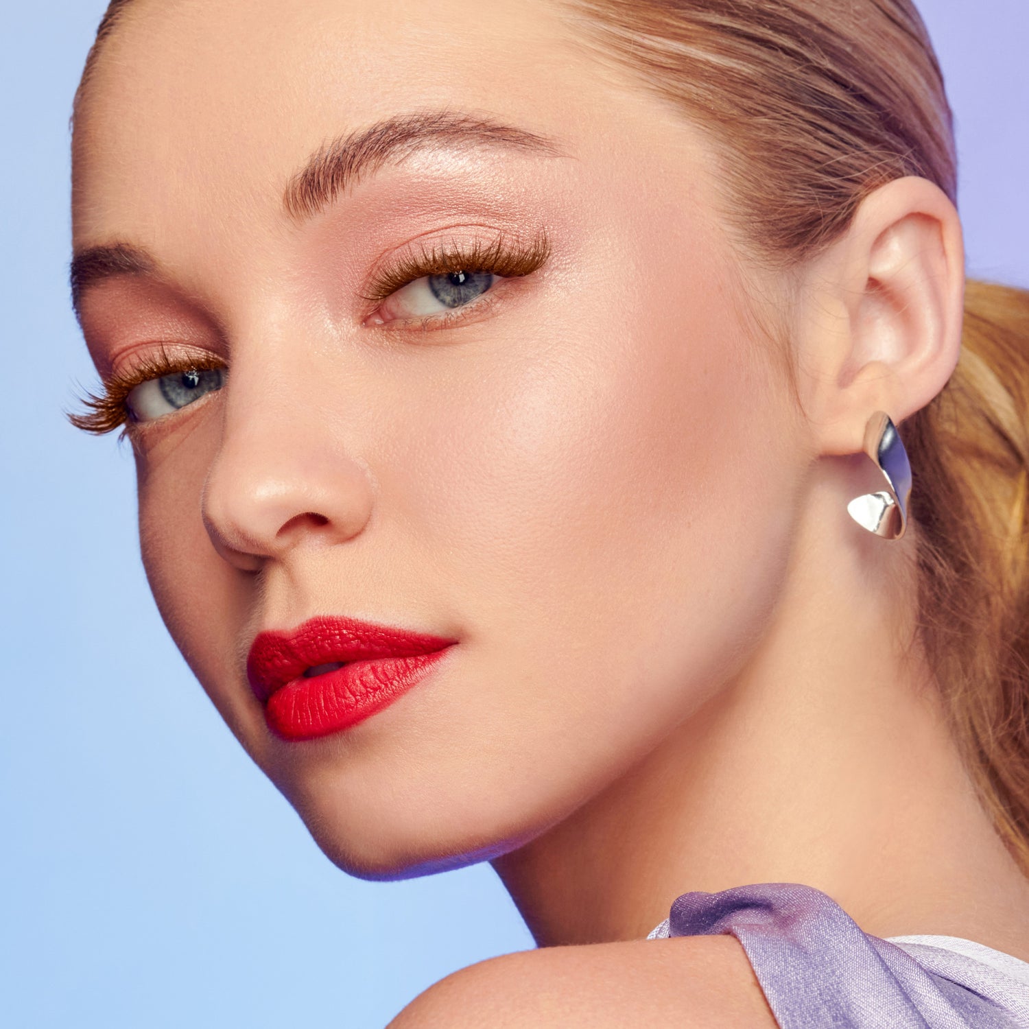 Close-up of a woman’s face showing brown wisps false eyelashes, light pink eyeshadow, bold red lipstick, silver earring, and a lilac strap over her shoulder against a light blue background.