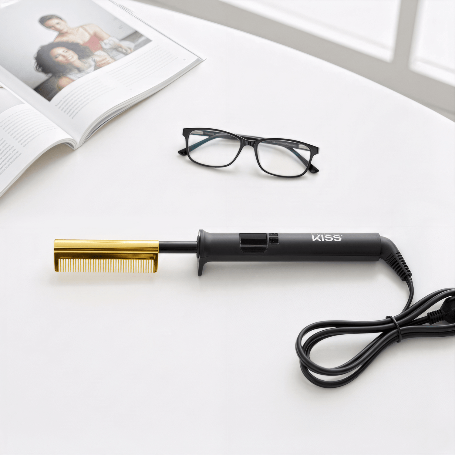 Black-handled KISS Professional Pressing Comb with a gold comb attachment, coiled power cord, resting on a white surface next to black-framed eyeglasses and an open magazine.