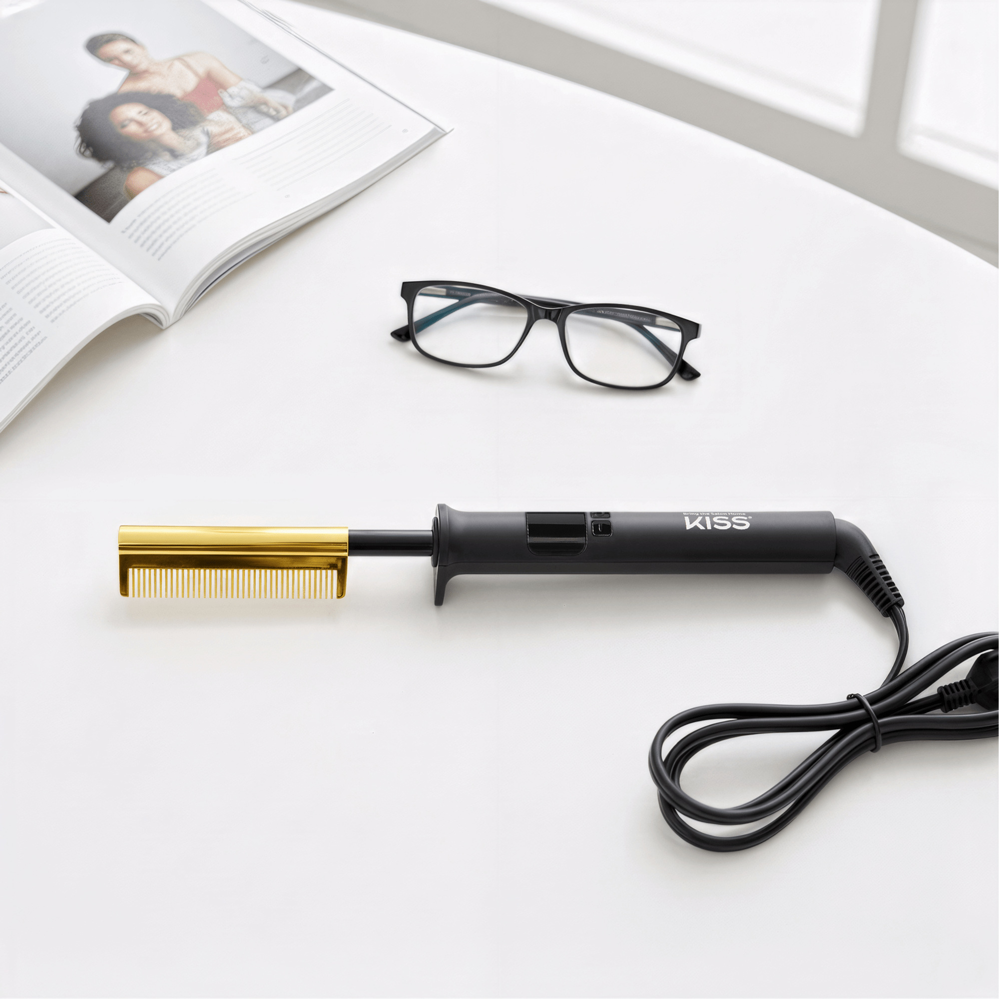 Black-handled KISS Professional Pressing Comb with a gold comb attachment, coiled power cord, resting on a white surface next to black-framed eyeglasses and an open magazine.