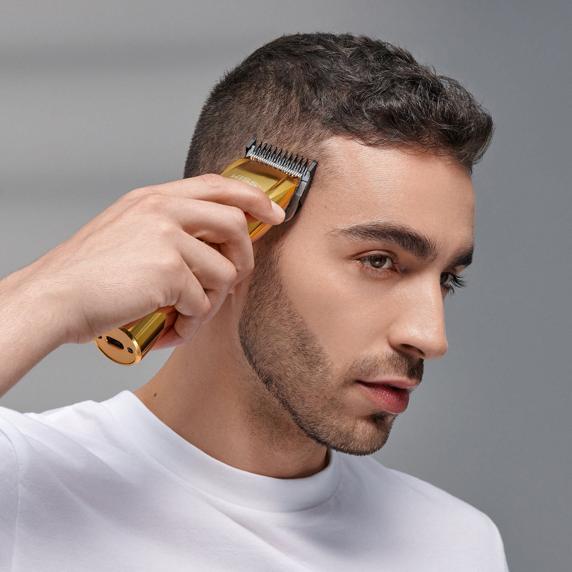 Man with short dark hair and trimmed beard using a gold cordless hair clipper on the side of his head, wearing a white shirt against a plain gray background.