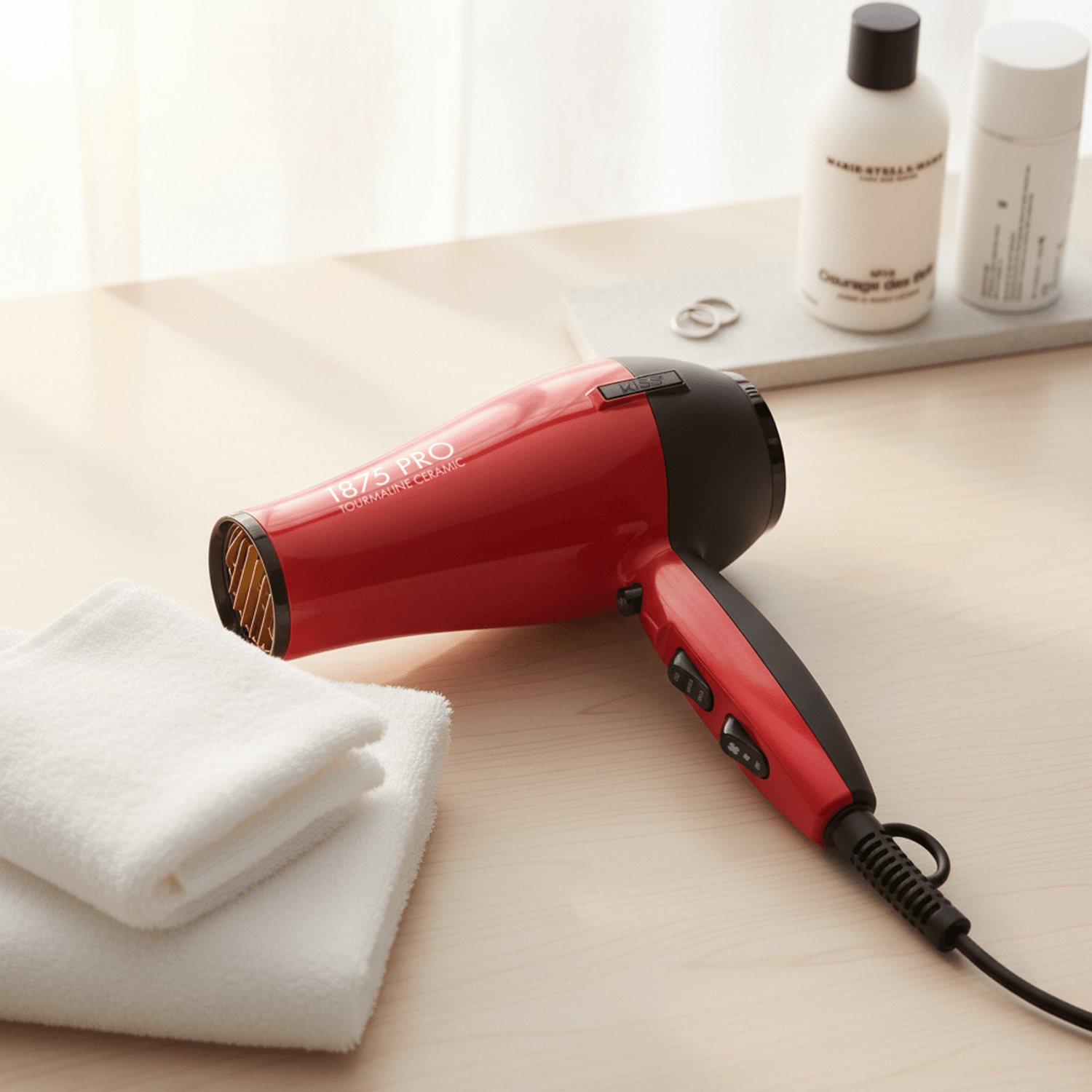 Red and black 1875 Pro Tourmaline Ceramic hair dryer resting on a light wood surface next to folded white towels, with bottles and rings blurred in the background.