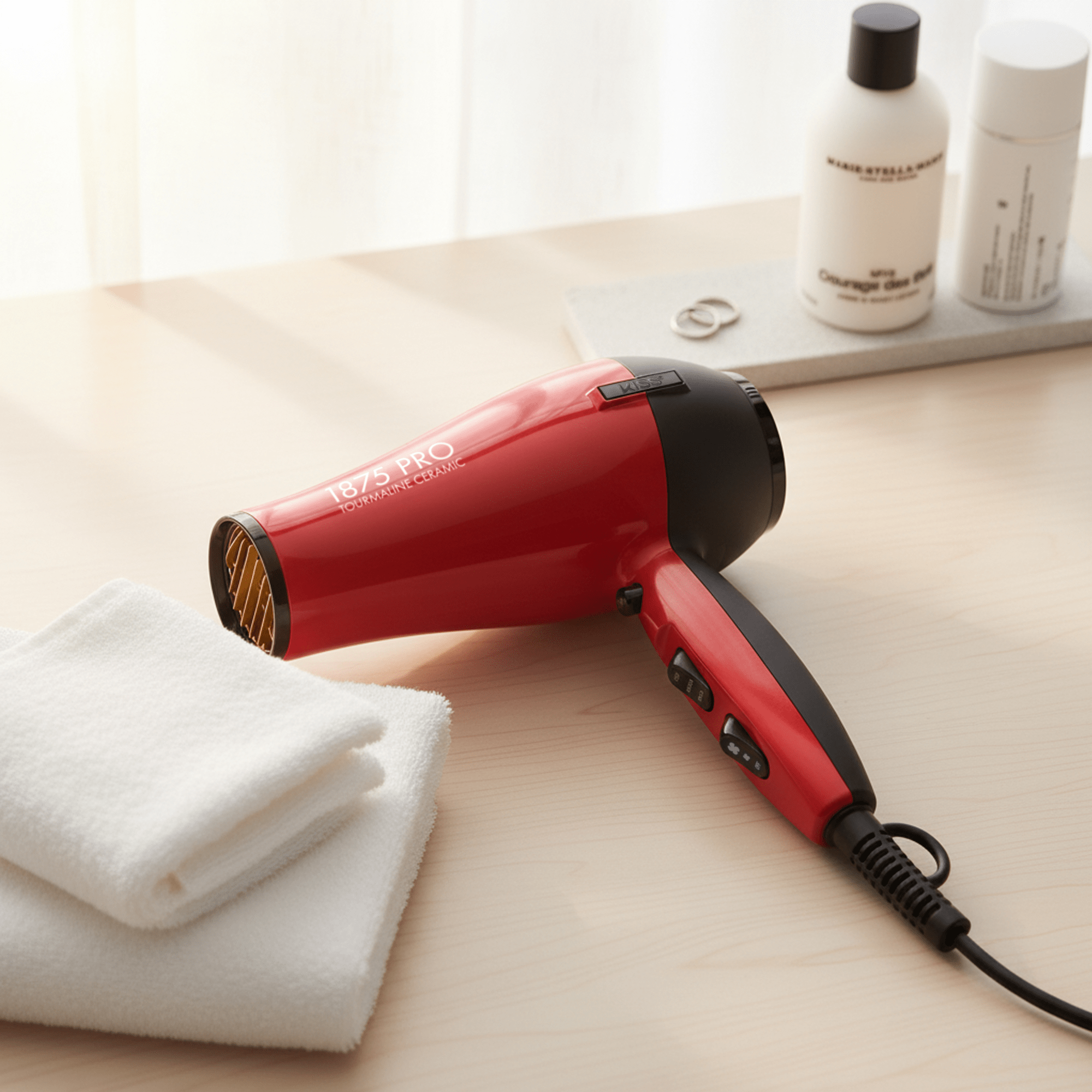 Red and black 1875 Pro Tourmaline Ceramic hair dryer resting on a light wood surface next to folded white towels, with bottles and rings blurred in the background.